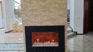 A modern fireplace with a stone facade and a glass front, situated in a spacious room with marble flooring and a staircase in the background.