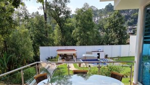A round glass table with four wicker chairs on a wooden deck, overlooking a lush garden and a white building with outdoor furniture.