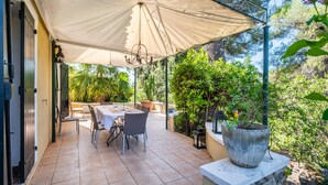 Een terras buiten met een tafel, stoelen, een grote parasol en potplanten.