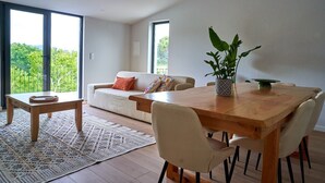 A modern living room with a wooden dining table, beige sofa, and a large window with a view of greenery.