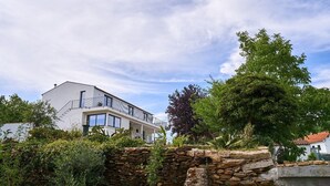 A modern two-story building with a balcony, surrounded by lush greenery and a stone wall.