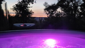 Un jacuzzi con luz púrpura, rodeado de árboles y un atardecer de fondo.