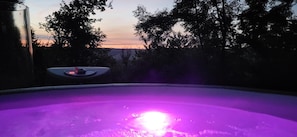 A hot tub with a purple light, surrounded by trees and a sunset in the background.