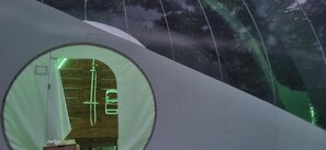 Een rond raam met houten binnenwerk, een hanglamp en uitzicht op een bos.