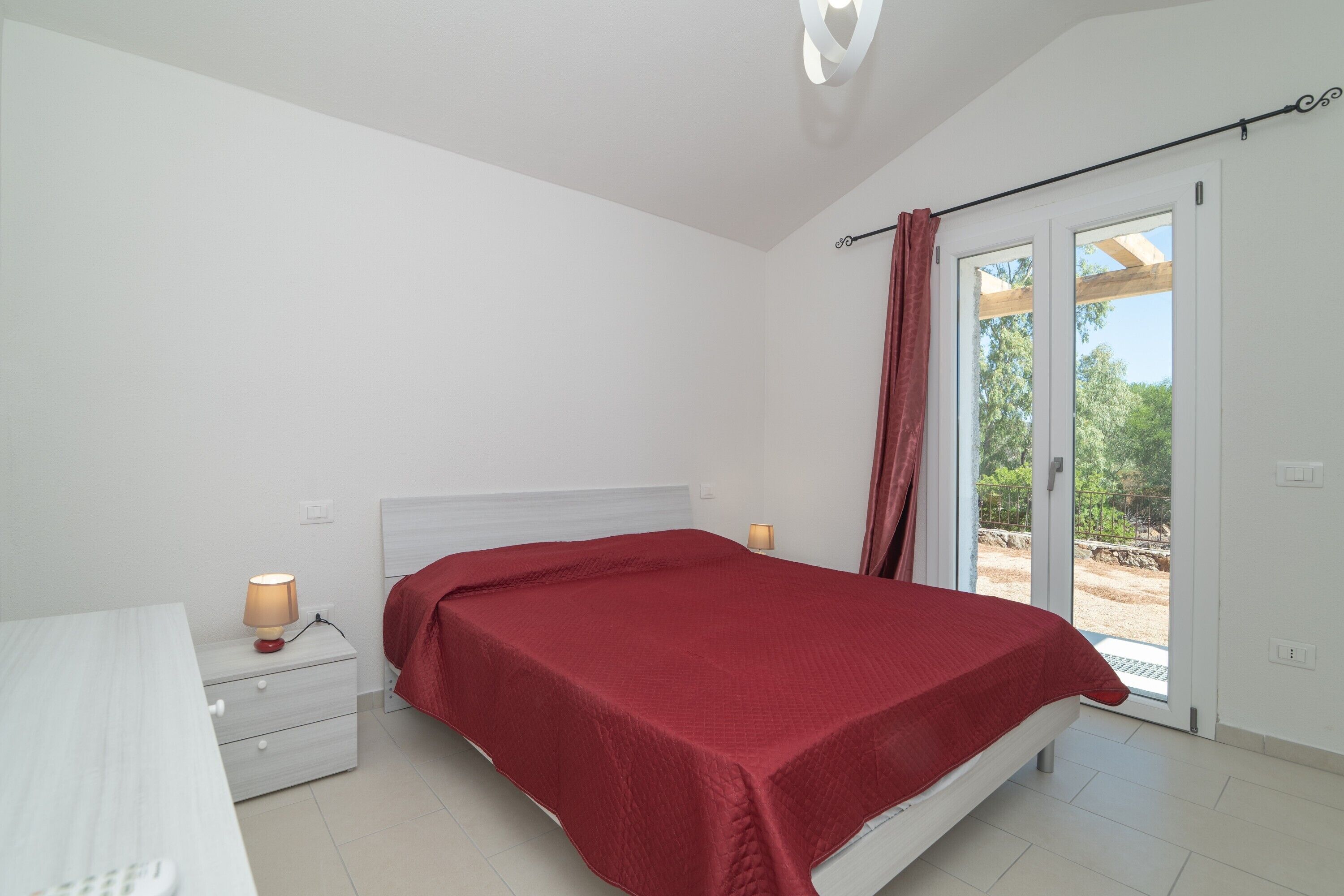 Un dormitorio con cama, mesitas de noche, una lámpara y un ventanal corredizo que da a un área exterior.