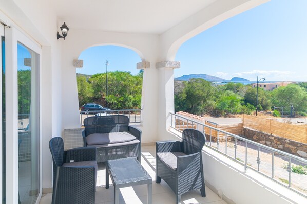 A balcony with wicker furniture, a view of greenery and distant buildings, and a clear sky.