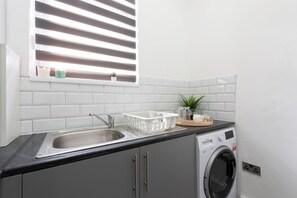 A kitchen with a sink, a washing machine, and a window with blinds. - The Grey Grove Pied-à-Terre at Stratford's Maryland Station (London)