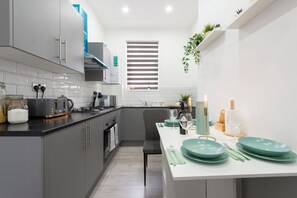 A modern kitchen with grey cabinets, a white countertop, and a dining area set with green plates and wine glasses. - The Grey Grove Pied-à-Terre at Stratford's Maryland Station (London)