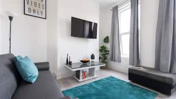 A modern living room with a grey sofa, a teal rug, a flat-screen TV, a small table with a plant, and a bench.
