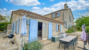 A stone building with a tiled roof, a patio with a table and chairs, and a grill outside.