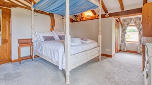 A four-poster bed with a canopy, a bedside table, and a dresser in a room with exposed wooden beams and a carpeted floor.