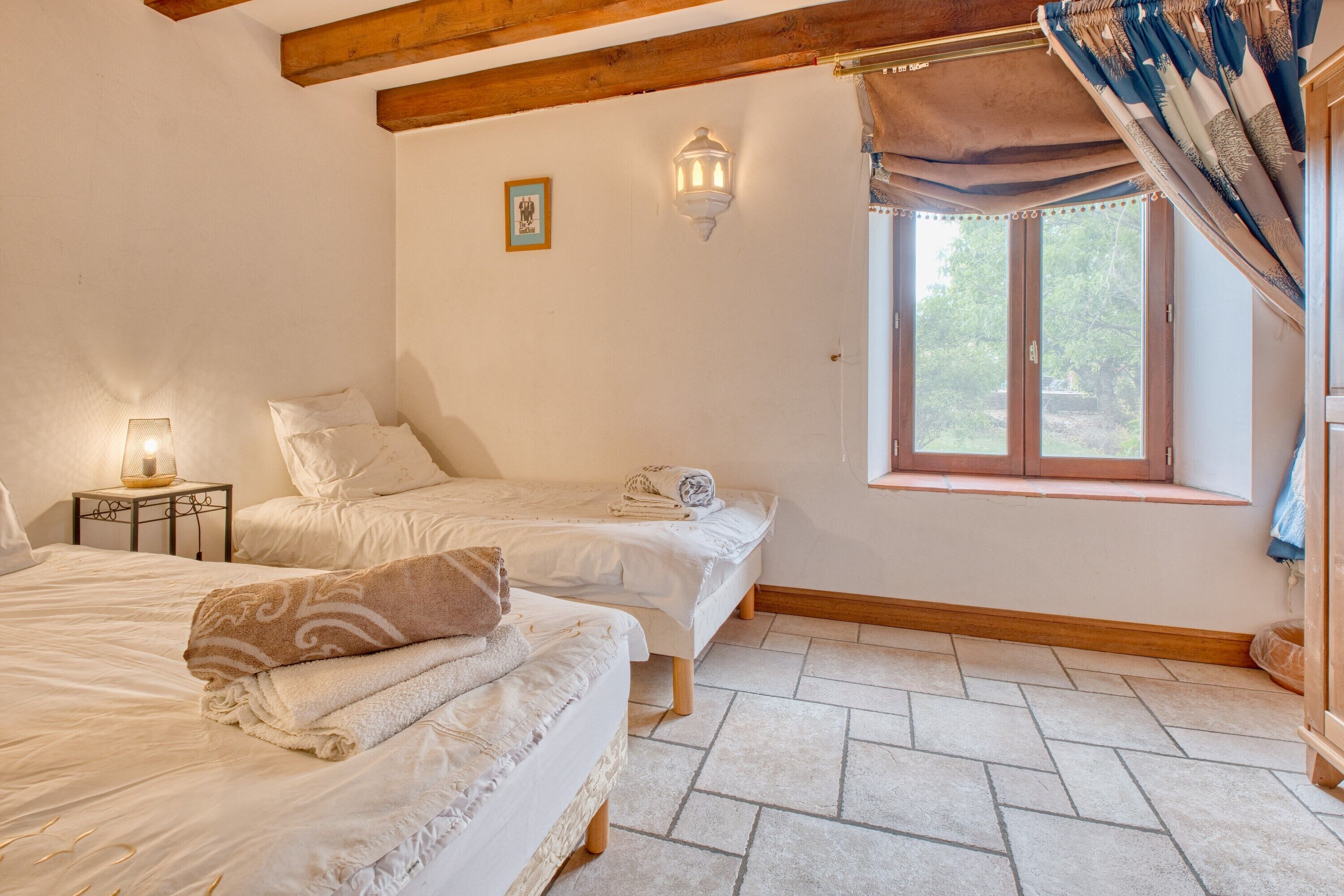Une chambre avec deux lits, une fenêtre avec des rideaux, une lampe fixée au mur et une petite table avec un lampadaire.