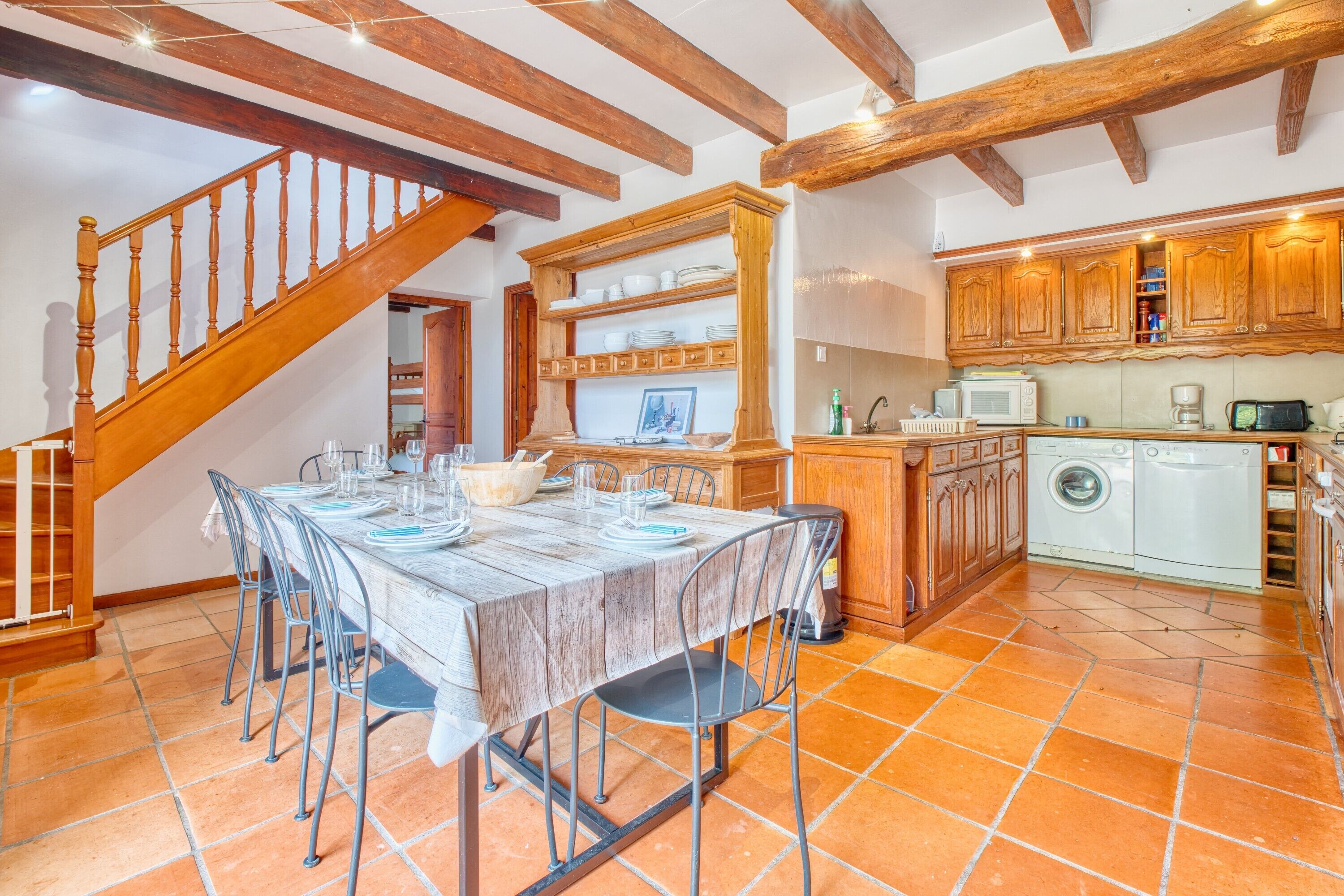 A dining area with a wooden table set for six, a kitchen with wooden cabinets, a washing machine, and a staircase.
