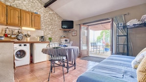 A compact kitchen with a washing machine, a dining area with a table and chairs, a bed with a blue cover, and a balcony with a view.