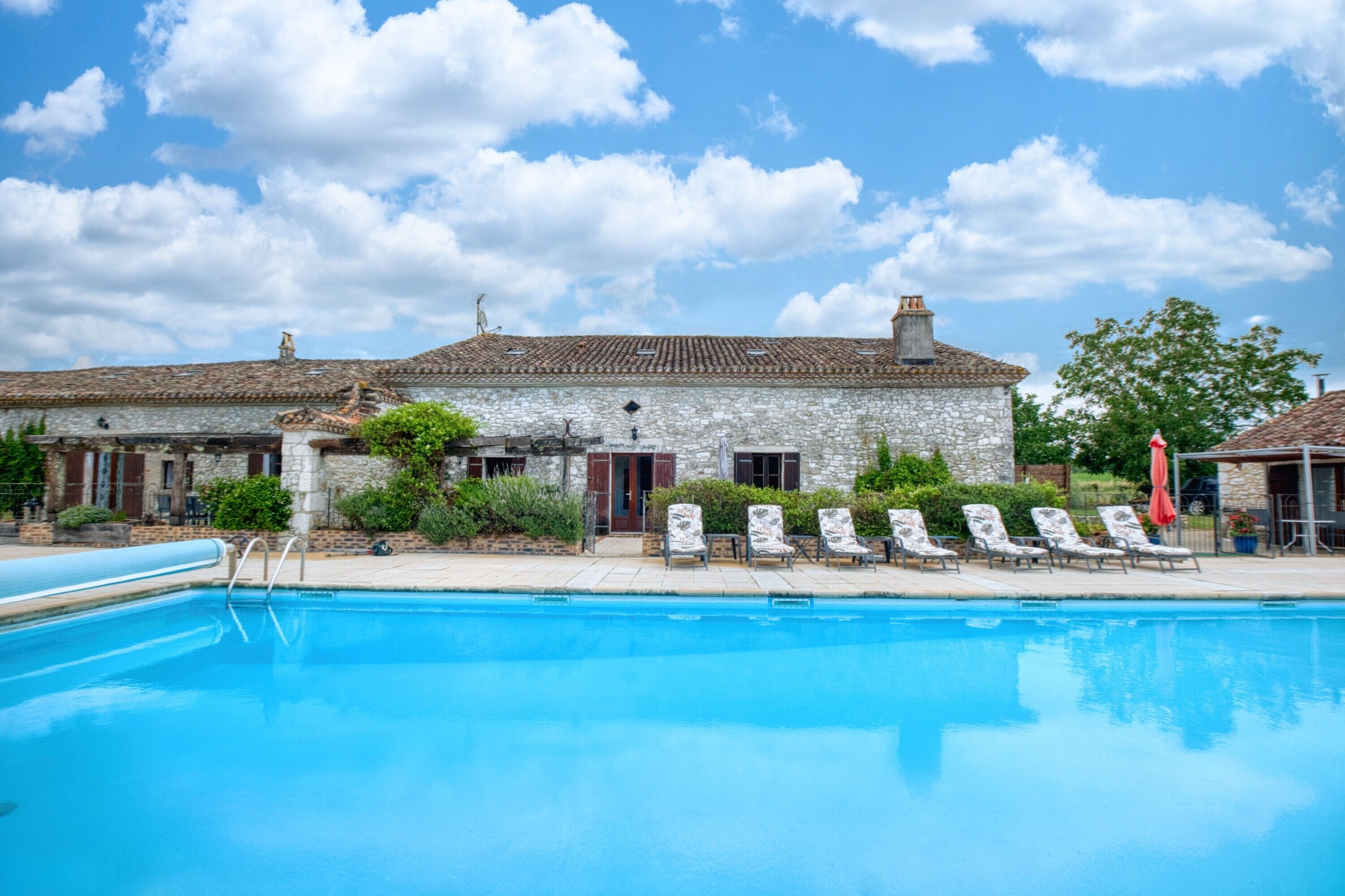 A swimming pool with lounge chairs, a stone building with a tiled roof, and a clear sky.