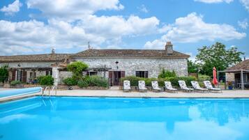 A swimming pool with lounge chairs, a stone building with a tiled roof, and a clear sky.