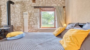 A bedroom with a stone wall, a bed with a quilted bedspread, pillows, a window with curtains, and a fireplace.
