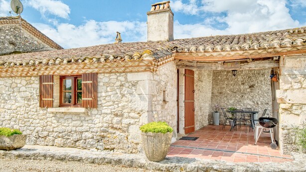 Un edificio de piedra con techo de tejas, una puerta y un patio con mobiliario exterior.