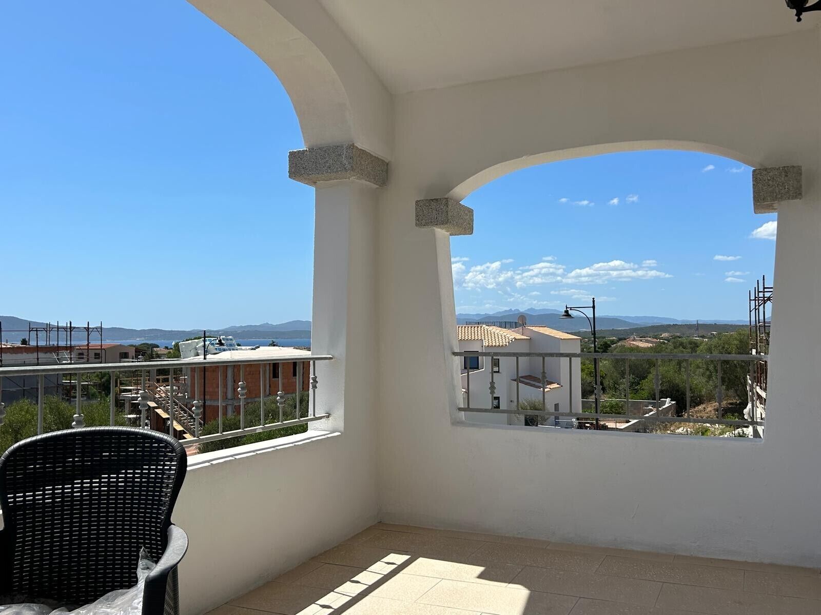 A balcony with a view of the sea and mountains, a wicker chair, and a tiled floor.