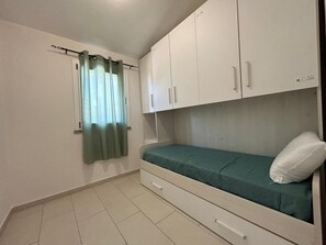 A small, single-bed room with a pull-out drawer, white cabinets, a window with green curtains, and a tiled floor.