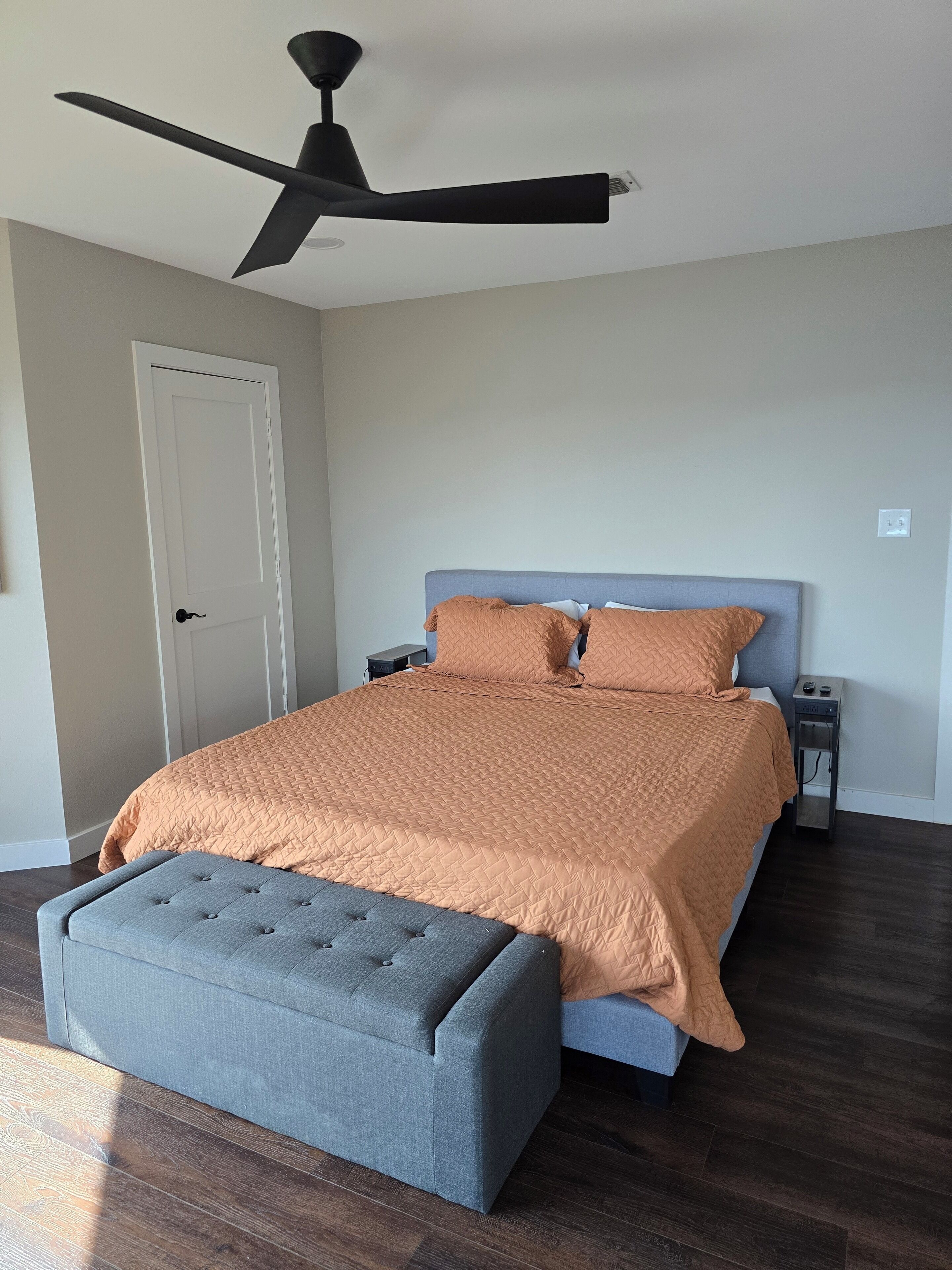 A bedroom with a bed, a grey upholstered bench, a ceiling fan, a nightstand, and a door.