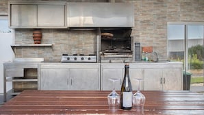 An outdoor kitchen with a brick wall, stainless steel cabinets, and a wooden table with wine bottles and glasses. - Villa 'Praia Azul Beach and Sun' with sea view, private garden, and Wi-Fi access (Silveira)