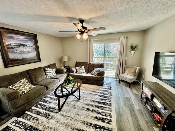 A living room with a sofa, armchair, and a coffee table. There is a ceiling fan, a TV, and a window with curtains.