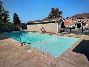 A swimming pool with a lifeguard chair and a lifebuoy, surrounded by a brick building and a fence.