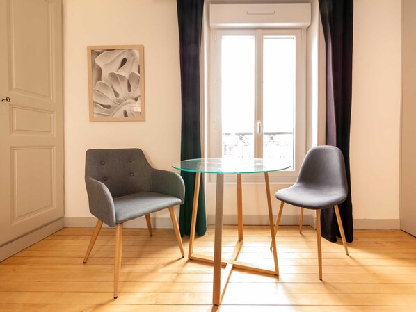 Une pièce avec une table en verre et deux chaises, une porte, une œuvre d’art abstraite encadrée et une fenêtre avec des rideaux.