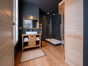 Une salle de bain moderne avec un meuble-lavabo en bois, un grand miroir, une douche et un mur recouvert de carreaux de couleur sombre.