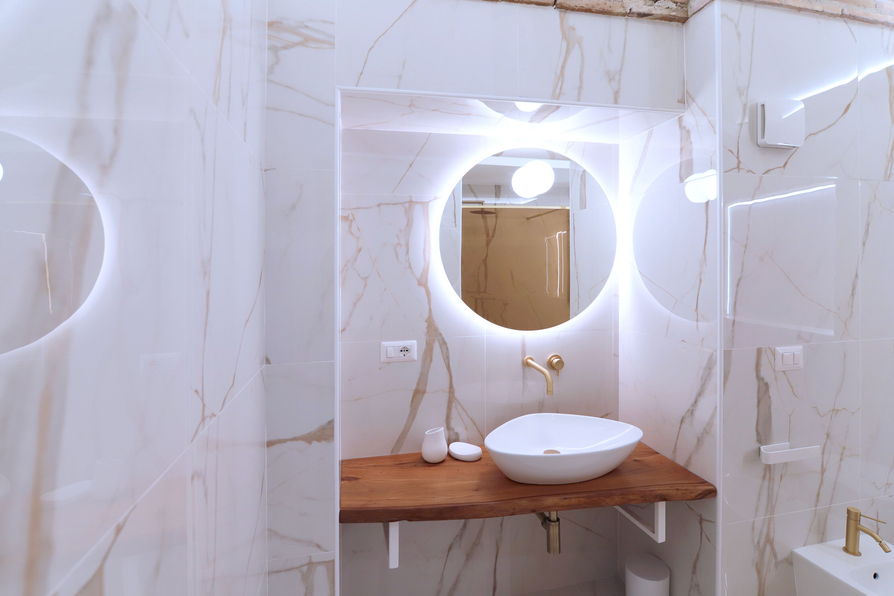 Une salle de bain dotée d’un lavabo en marbre blanc, d’un plan de toilette en bois et d’un miroir rond avec éclairage intégré.