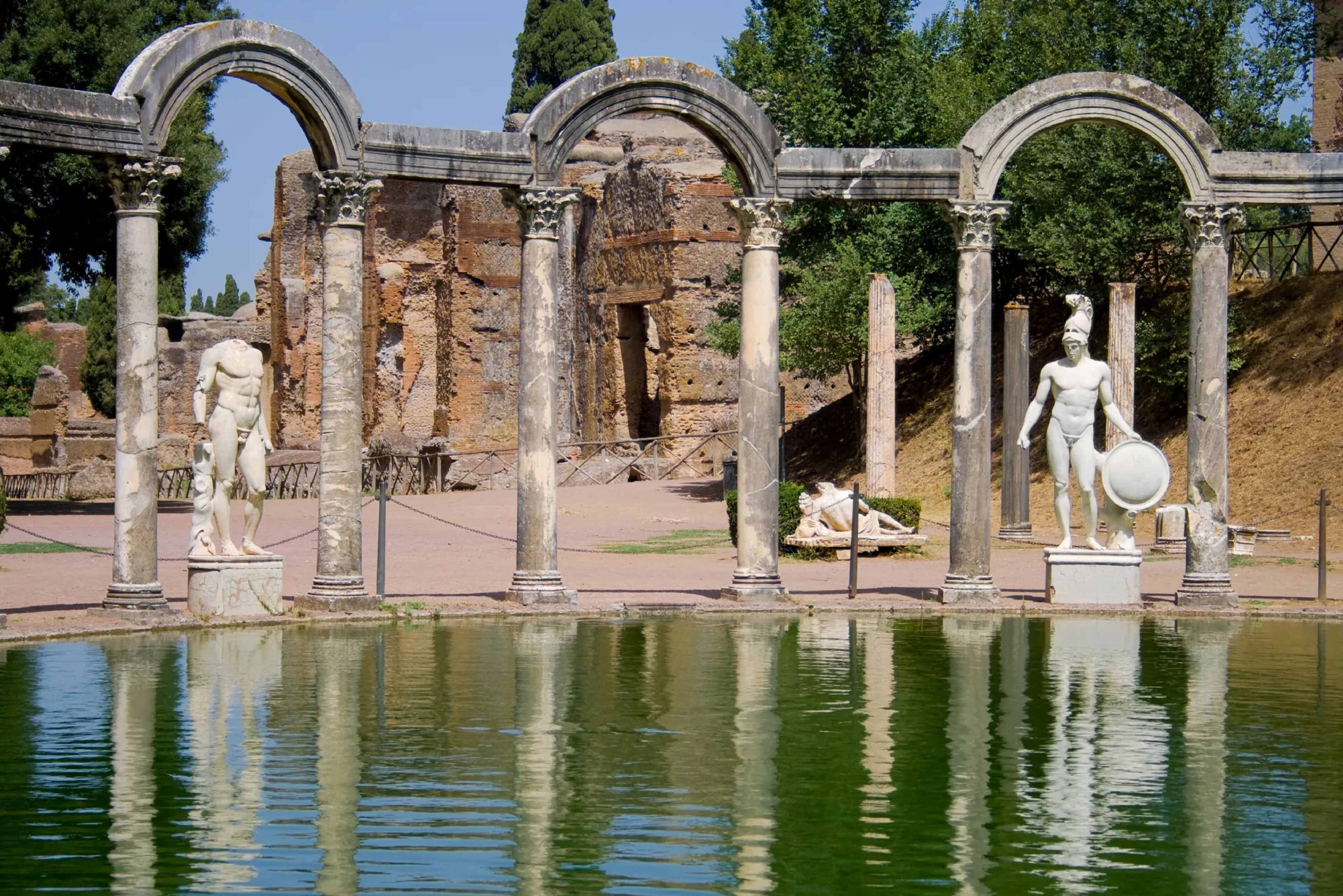 Rovine antiche con colonne e statue accanto a uno specchio d'acqua.