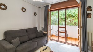 Una sala moderna con un sofá, un control remoto sobre una mesita redonda y un ventanal corredero que da a un área exterior con una mesa y sillas.