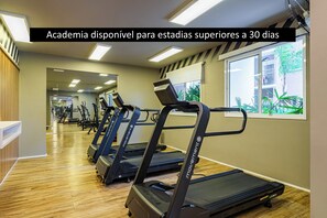 A gym with treadmills and exercise equipment, a reception desk, and large windows with a view of the outside. - Laguna 1412 - Moderno e Funcional (Sao Paulo)