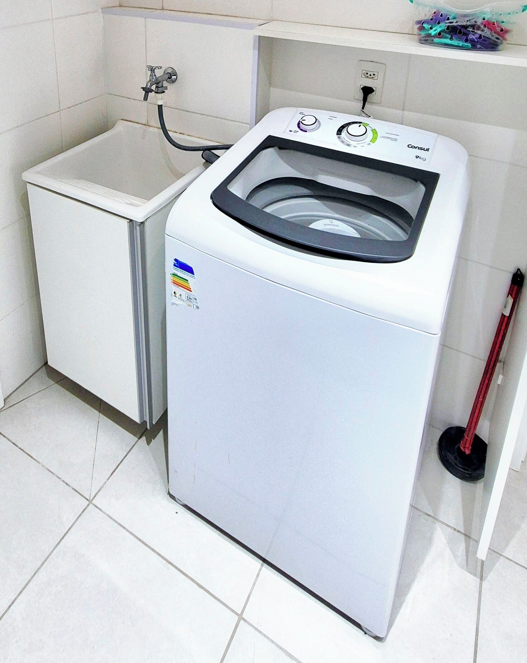 Een compacte, witte wasmachine met een bedieningspaneel en een transparante deur, naast een witte wasbak en een rode dweil in een betegelde badkamer.