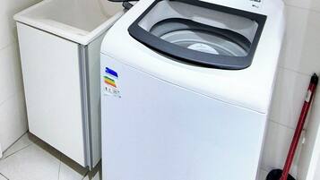 Een compacte, witte wasmachine met een bedieningspaneel en een transparante deur, naast een witte wasbak en een rode dweil in een betegelde badkamer.