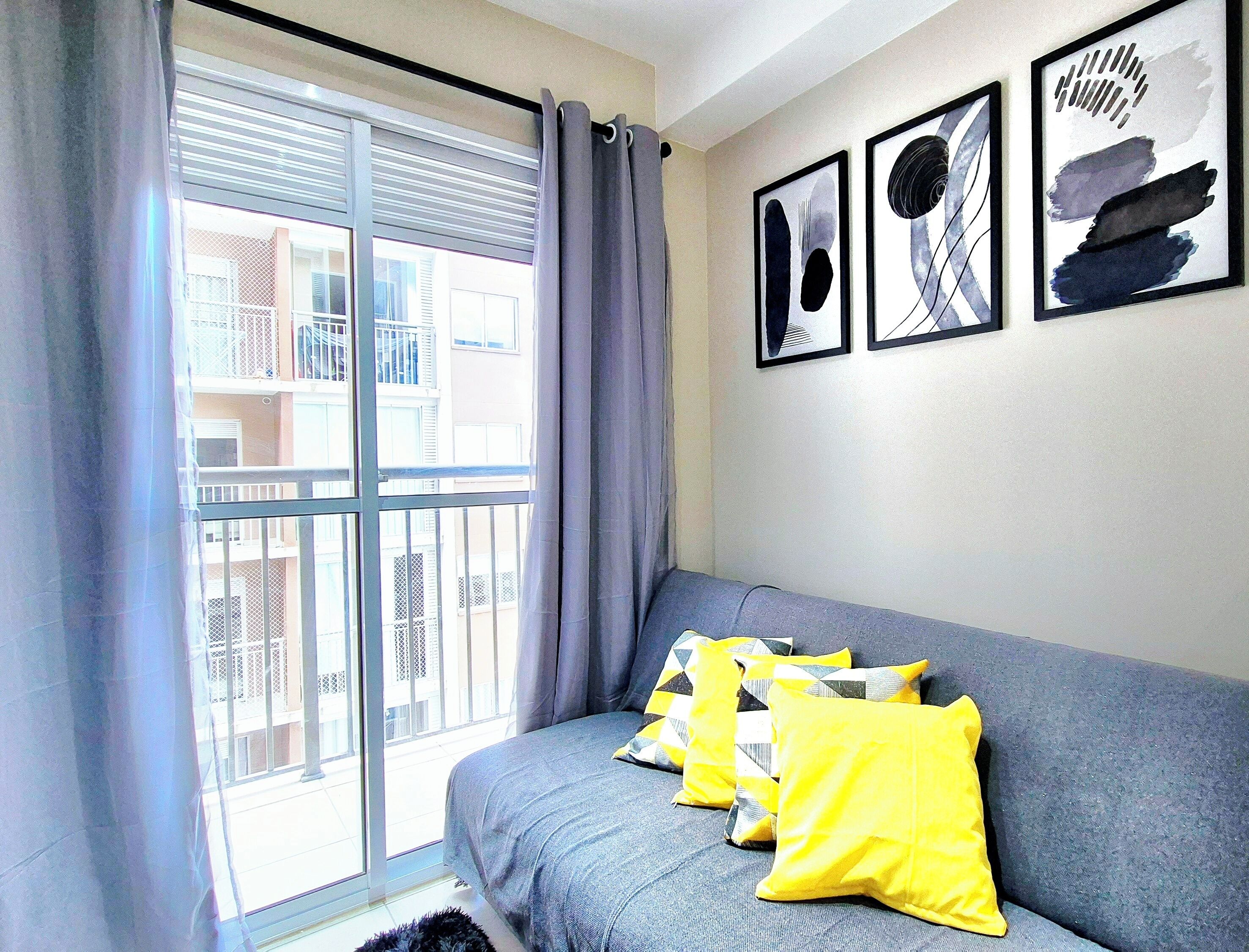 A room with a grey sofa, yellow pillows, a large window with curtains, and three framed artworks on the wall.