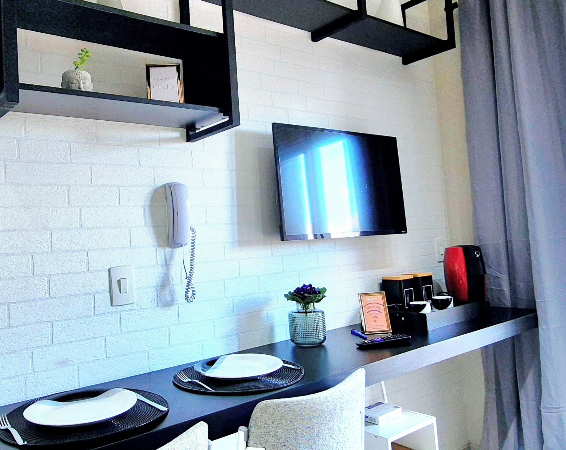A modern hotel room with a white brick wall, a mounted flat-screen TV, a telephone, a small plant, a framed certificate, and a dining area with a table set for two.