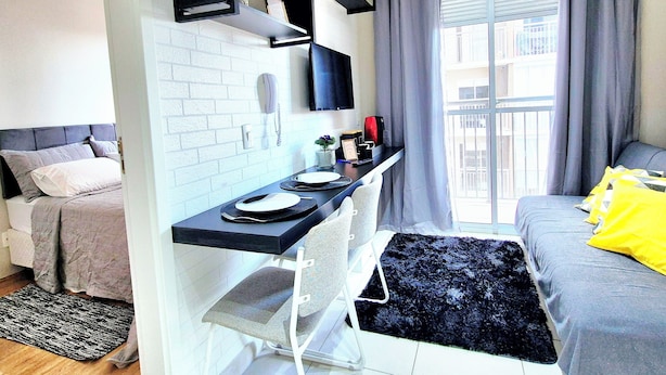 A compact hotel room with a bed, a small kitchenette, a dining area, and a balcony with curtains.