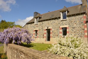 A stone building with a sloped roof, a small porch, and a garden with purple and white flowering plants.