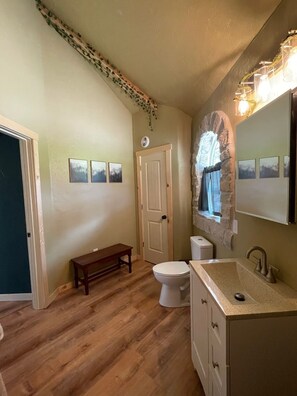 Een badkamer met een houten vloer, een bankje, toilet, wastafel met spiegel en ingekaderde foto's aan de muur.
