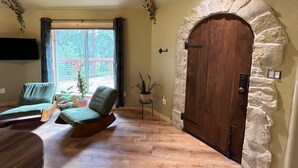 A room with a stone arch doorway, two green cushioned chairs, a wooden table, a potted plant, and a window with a view of a deck.