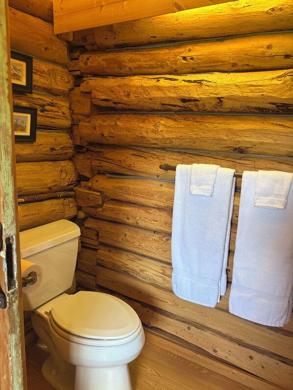 Une salle de bain rustique, aménagée dans une cabane en bois, avec des toilettes et deux serviettes suspendues à un porte-serviettes.