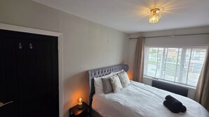 A bedroom with a large bed, a bedside table with a lamp, a window with blinds, and a door.
