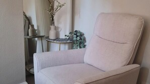 A beige armchair in a corner of a room with a small round table and a tall white vase with branches.