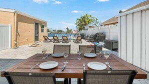 A patio with a wooden table set for dining, surrounded by lounge chairs and a barbecue grill.