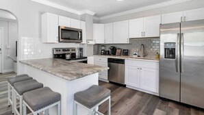 A modern kitchen with white cabinets, stainless steel appliances, a marble countertop, and bar stools.