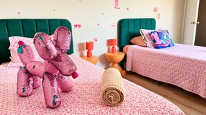 Un dormitori amb dos llits, un os de peluix amb pedreria rosa, una tovallola enrotllada i una paret amb pegatines decoratives.