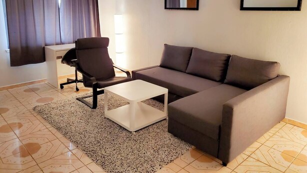Una sala de estar moderna con un sofá marrón, una mesa de centro blanca, una silla de oficina negra y un pequeño escritorio blanco. Hay dos espejos en la pared y una lámpara en el techo.