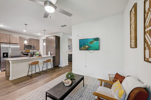 A modern kitchen with a central island, bar stools, and a flat-screen TV mounted on the wall. - Luxe Poolside Condo, King Bed, Near Mayo Clinic, St Johns Town Center & Beaches (Jacksonville)